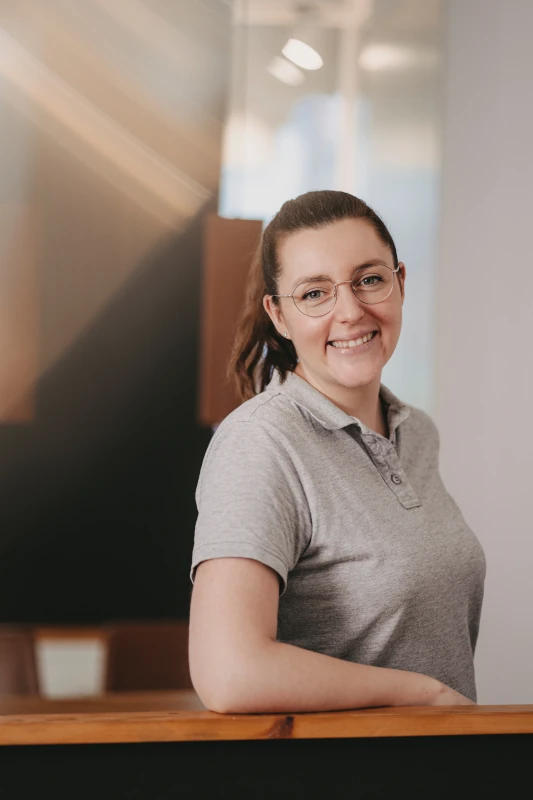 Freundliche Frau mit Brille und grauem Poloshirt lächelt an Empfangstresen im Büro.