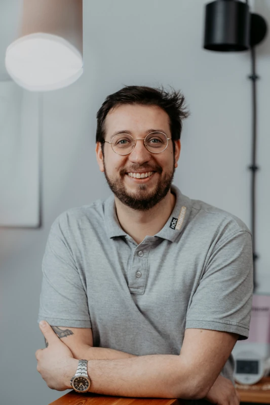 Mann mit Brille und Bart lächelt freundlich in modernem Büro mit grauer Wand.